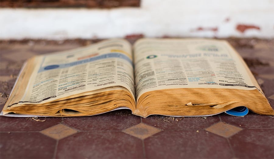 A weathered, yellowed phone book lies open on a tiled floor with frayed pages, suggesting age and frequent use.
