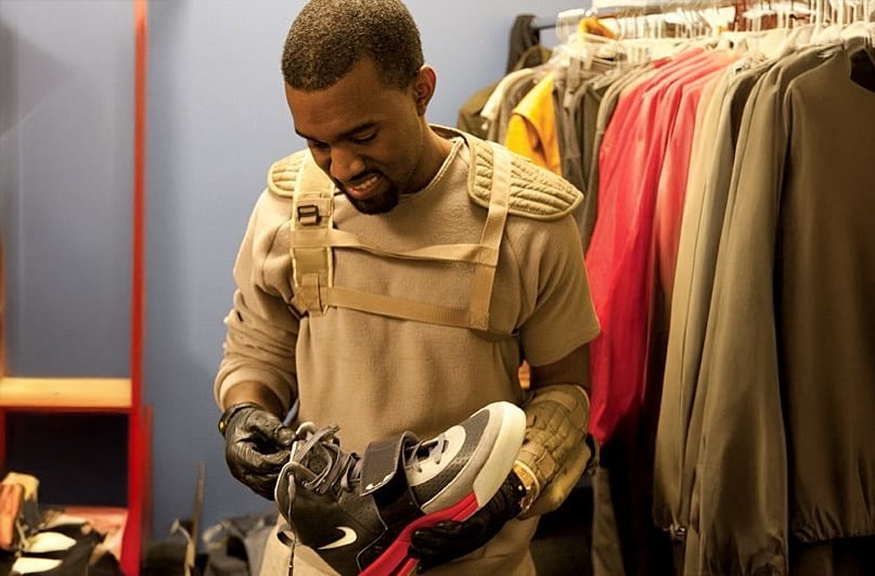 A man wearing a tan outfit and black gloves holds and looks at a black and pink Nike sneaker while standing in a closet with jackets hanging on a rack.