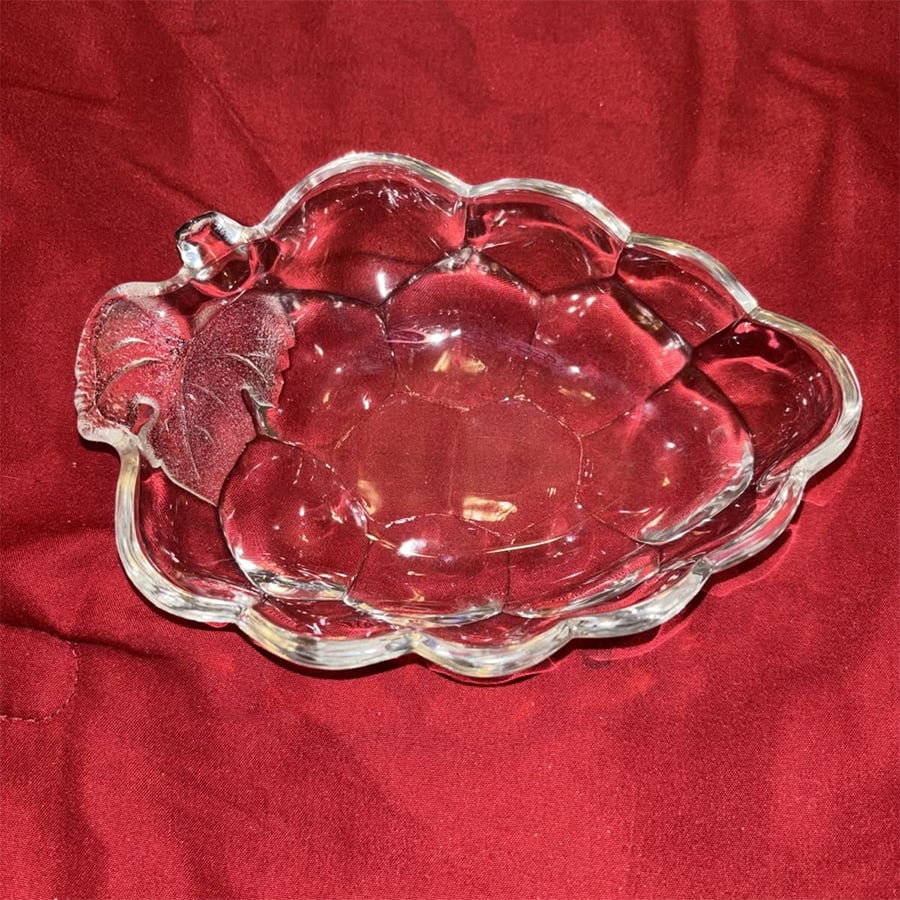 A clear glass dish shaped like a leaf, with textured detailing, sits on a red fabric background.