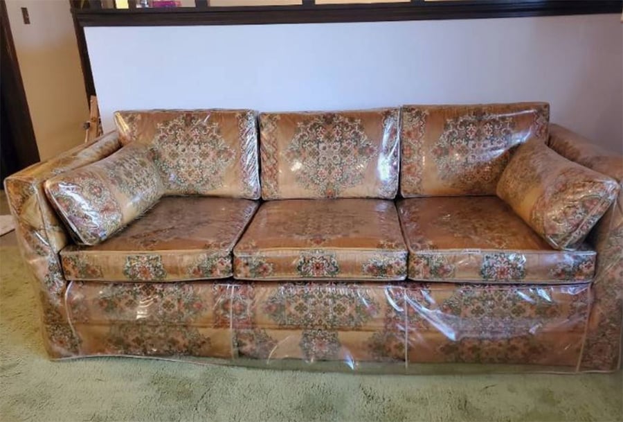 A vintage floral-patterned couch covered in clear plastic sits on green carpet against a light-colored wall.