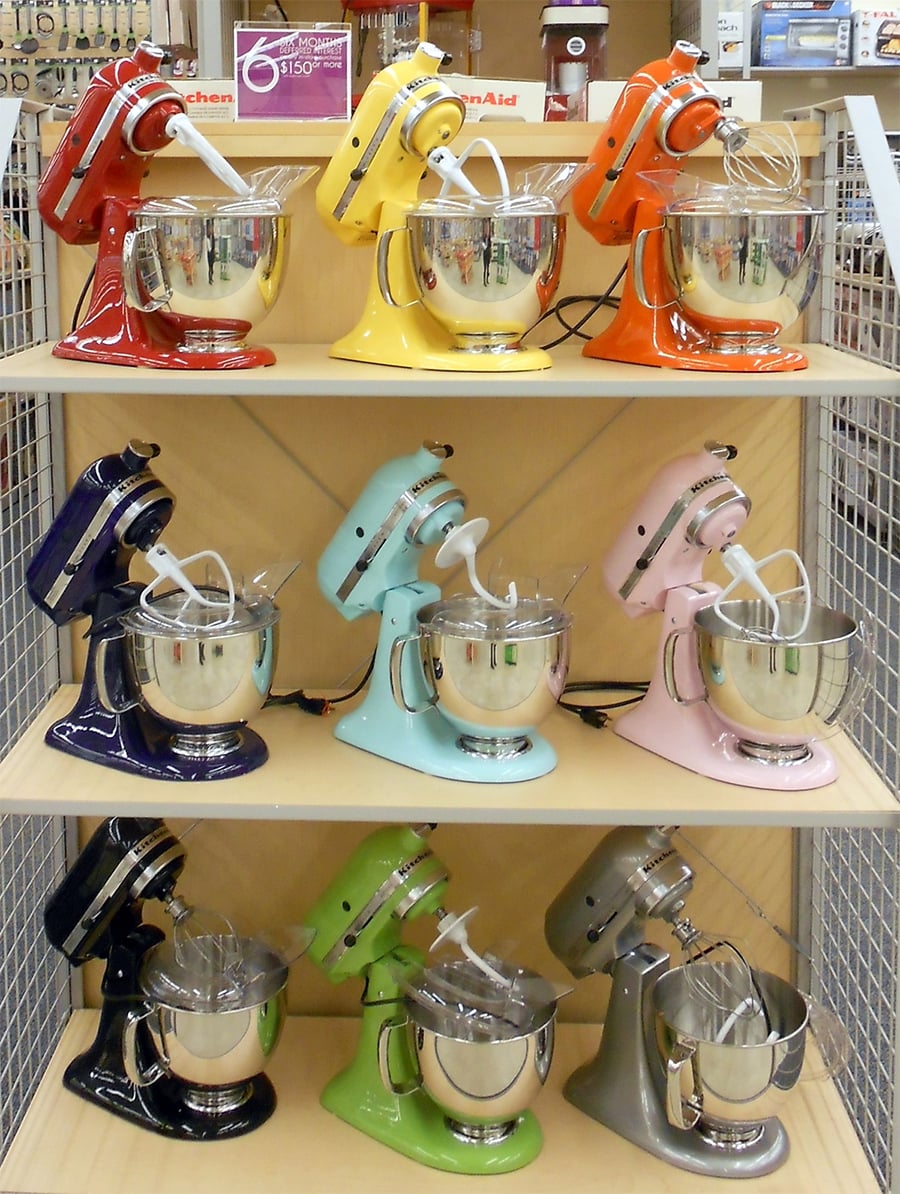 Nine colorful stand mixers in red, yellow, orange, black, light blue, pink, dark blue, green, and silver are displayed on wooden shelves in a store. Each mixer has a shiny metal bowl and beater attachment.