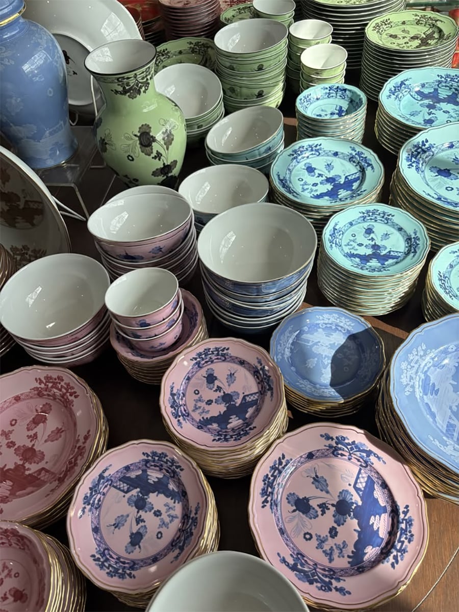 Stacks of colorful plates, bowls, and vases with intricate patterns in pink, blue, green, and white are arranged on a table, with sunlight casting soft shadows across the ceramics.