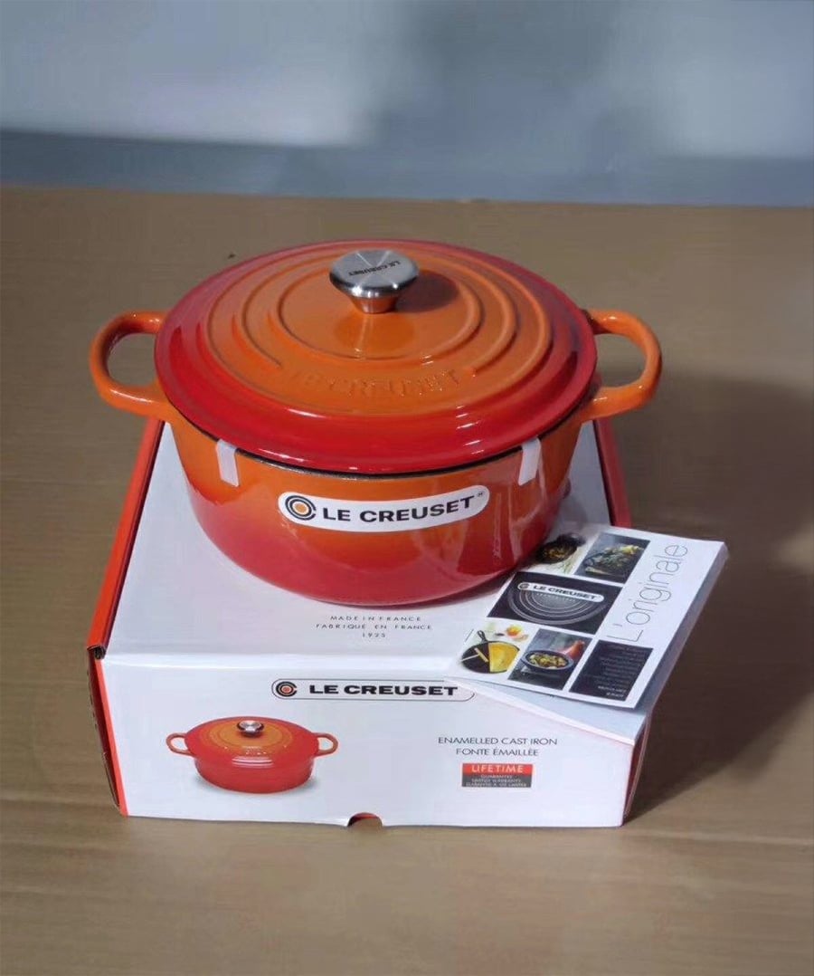 An orange Le Creuset enameled cast iron Dutch oven sits on its box, with product booklet beside it, on a wooden surface.