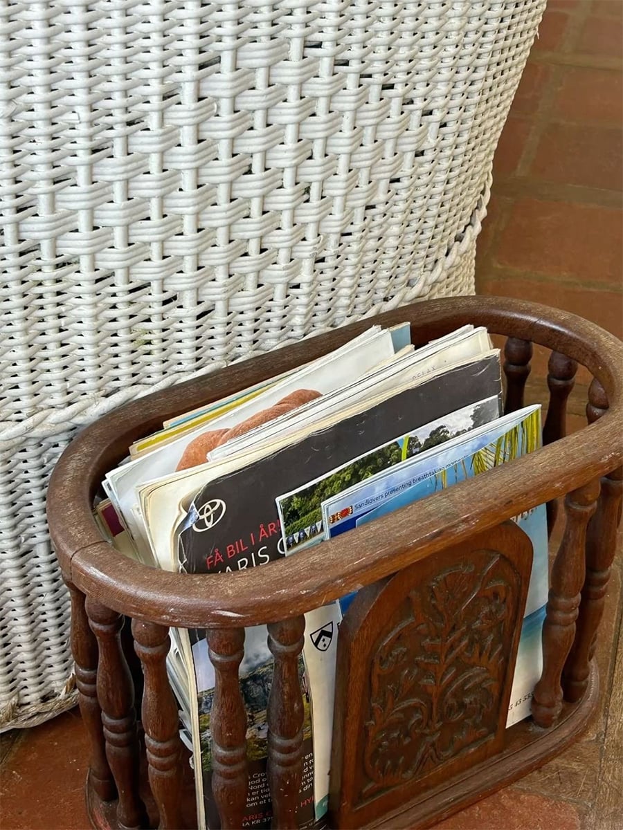 A wooden magazine rack filled with various magazines and papers sits on a tile floor beside a white woven basket.
