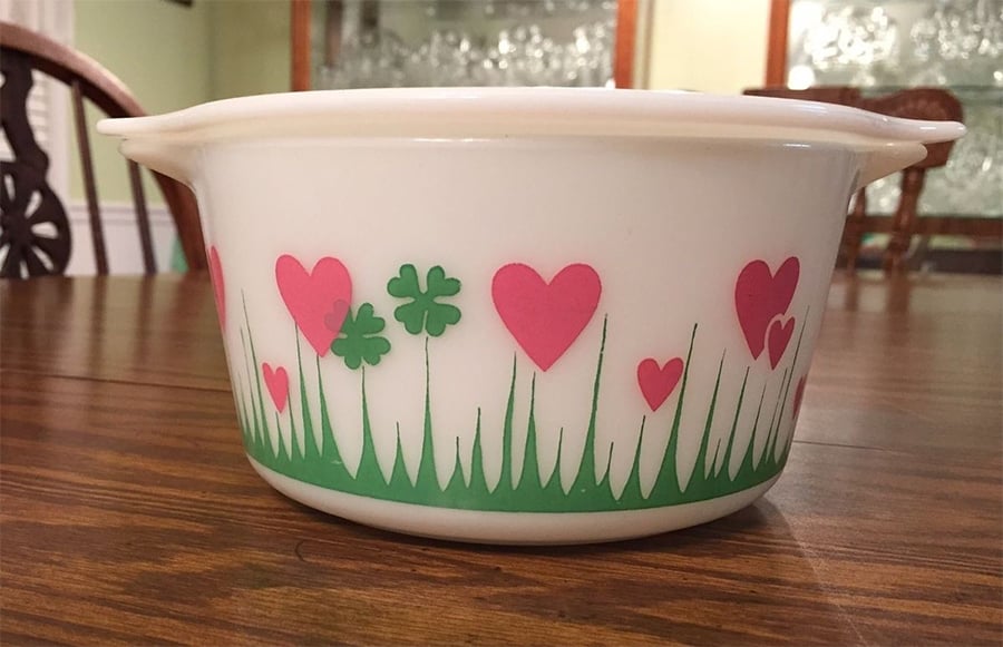 A white casserole dish decorated with green grass, green clovers, and pink hearts sits on a wooden table in a dining room with chairs and a glass-front cabinet in the background.