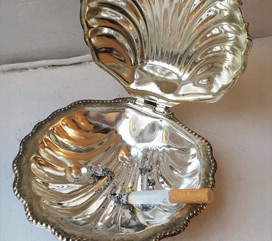 A gold, clam shell-shaped box is used as an ashtray, holding a cigarette and some ashes inside.