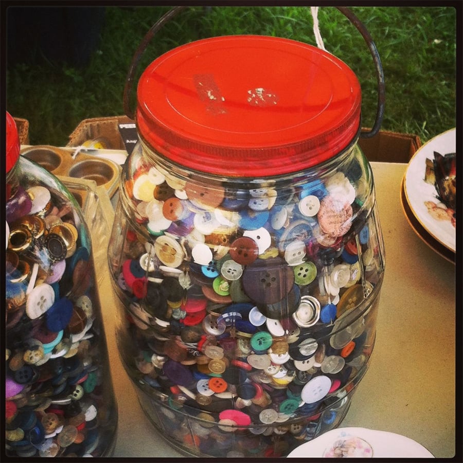 A large clear jar with a red lid is filled with assorted colorful buttons, sitting on a table next to other jars and plates.