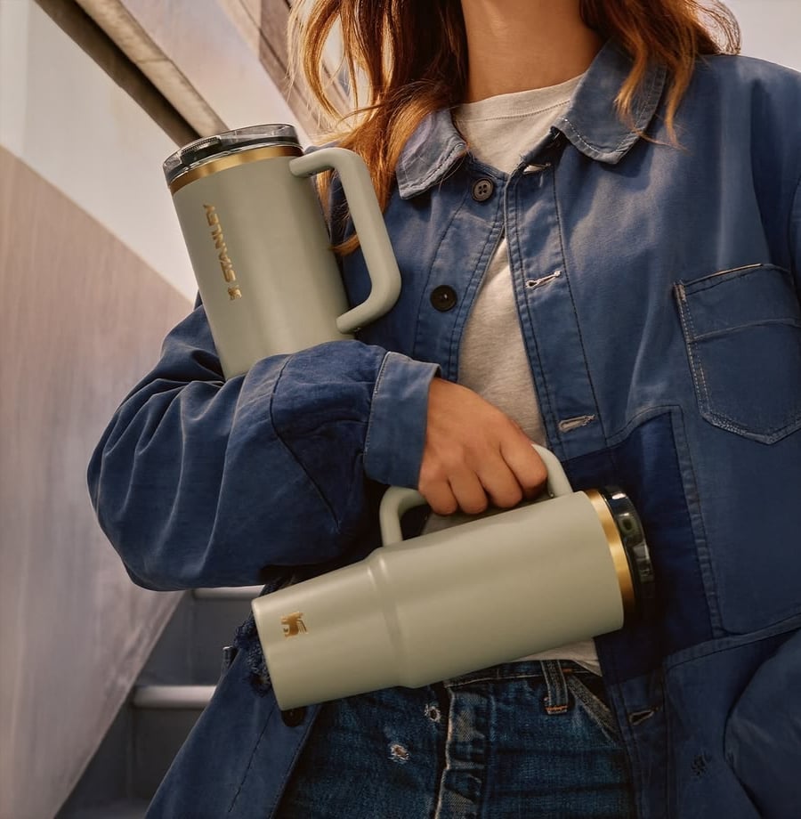 A person in a blue jacket holds two light-colored travel mugs with handles and black lids, standing on a staircase.