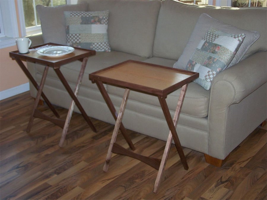A beige sofa with two patterned pillows sits behind two wooden TV trays; one tray holds a plate and cup, the other is empty. The floor is wood, and windows are in the background.