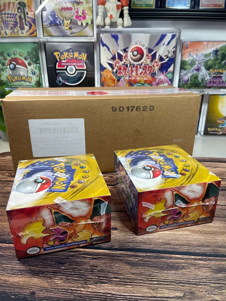 Two sealed Pokémon trading card game booster boxes with yellow and red designs sit on a wooden table in front of a brown shipping box. Pokémon posters are displayed in the background.