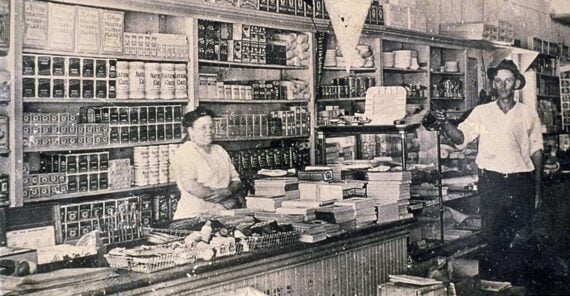 Black and white photo of an old-fashioned grocery store with shelves stocked with cans and goods. A woman stands behind the counter and a man stands nearby, both surrounded by various products.