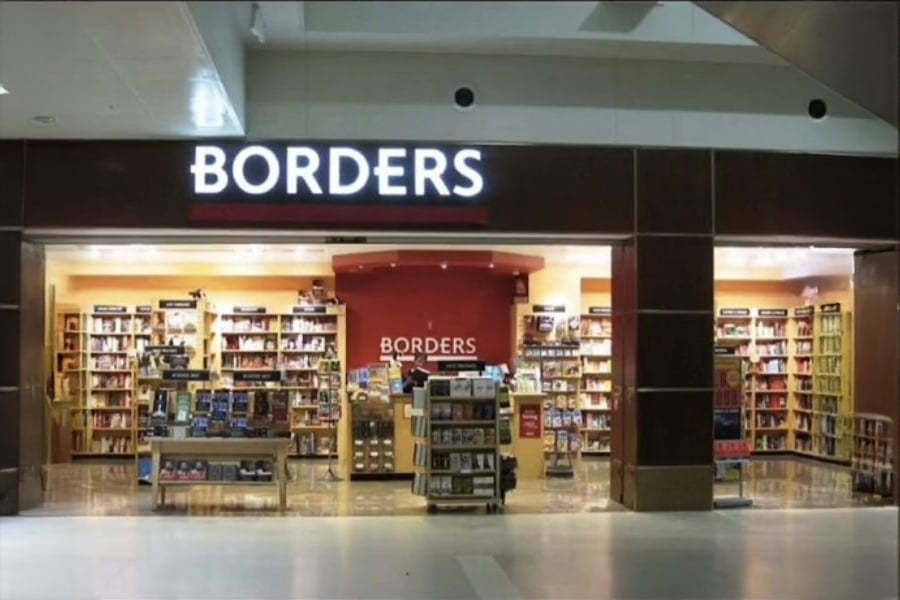 A Borders bookstore with illuminated sign, filled with shelves of books and displays, seen from outside in a shopping mall.