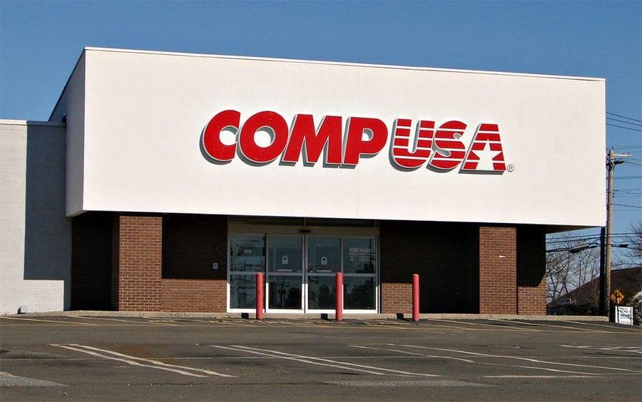 A closed CompUSA store with a large red and white sign above the entrance, empty parking spaces, glass doors, and three red bollards in front.