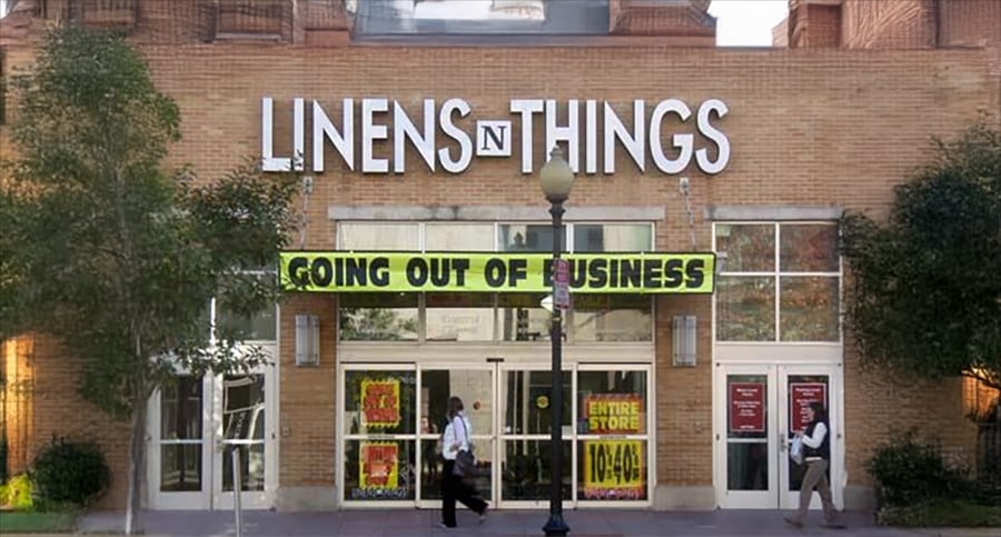 A Linens 'n Things store with a large yellow "Going Out of Business" banner above the entrance. Two people stand near the doors, and sale signs are visible in the windows.