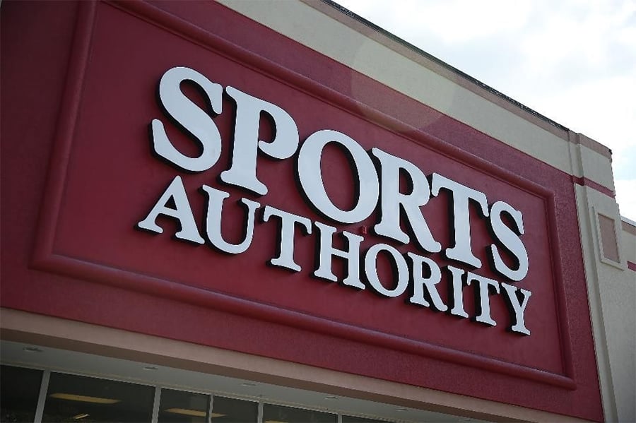 A large red and white Sports Authority store sign is mounted on the front exterior of a building under a partly cloudy sky.
