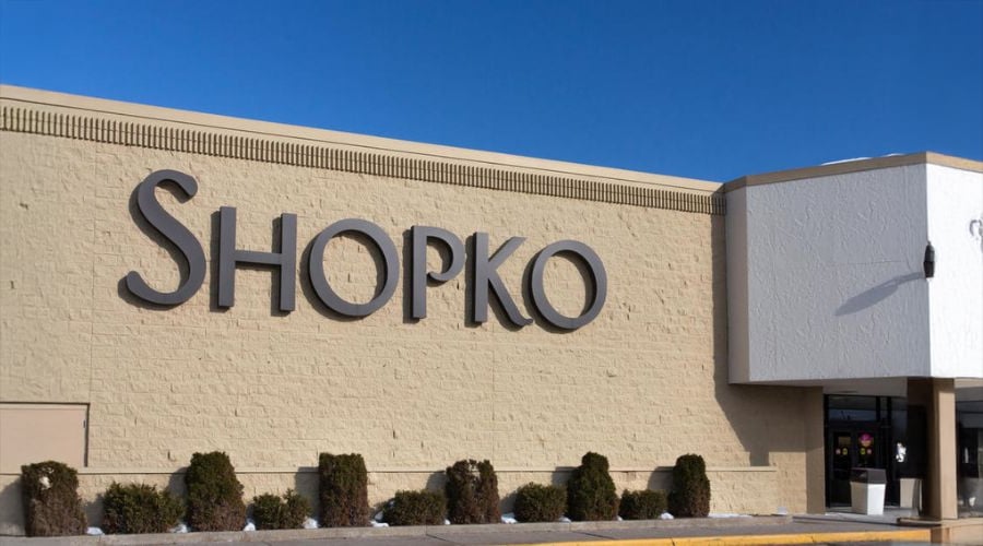 The exterior of a Shopko retail store, featuring a large "SHOPKO" sign on a tan brick wall, with bushes lined up outside and a clear blue sky above.