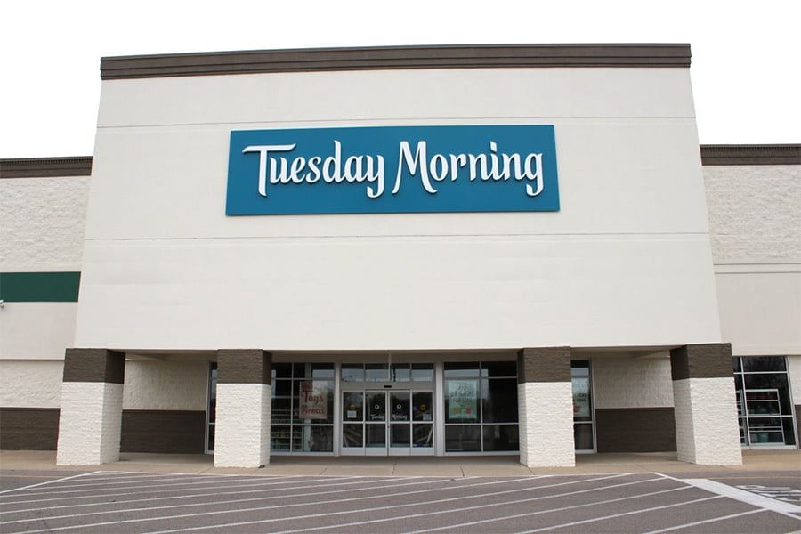 A Tuesday Morning store with a teal sign and glass doors sits in an empty parking lot, its white and brown facade visible under a cloudy sky.