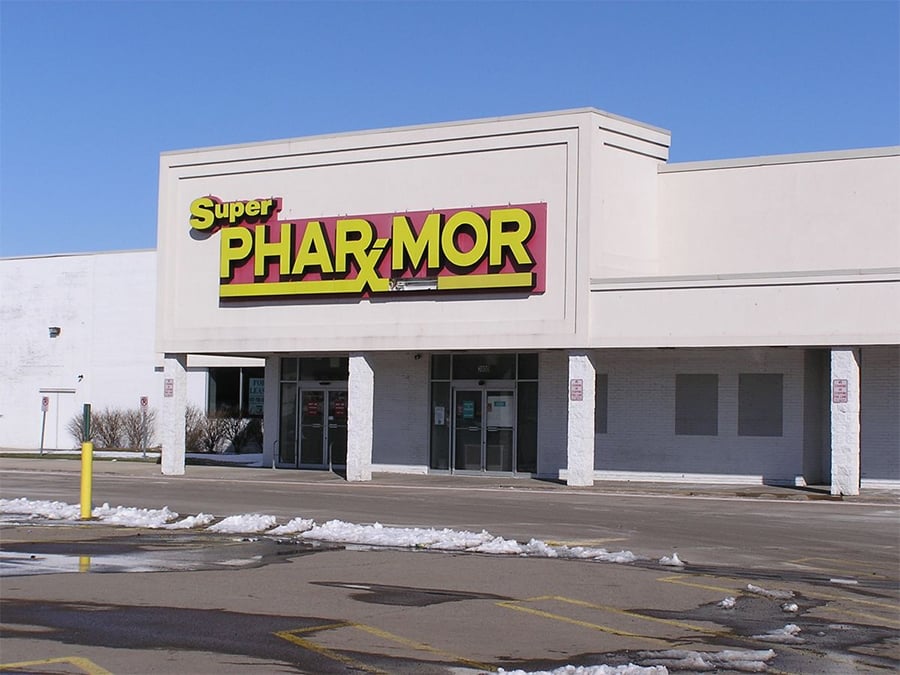 A large, white Super Pharrmor store with bold yellow and red signage on the building, seen from the mostly empty parking lot with some snow on the ground under a clear blue sky.