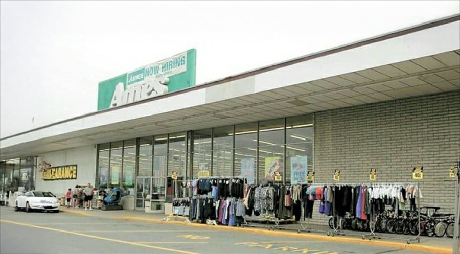 The exterior of an Ames department store, with clothing racks and bicycles displayed on the sidewalk, a clearance sign, and a white car parked outside.