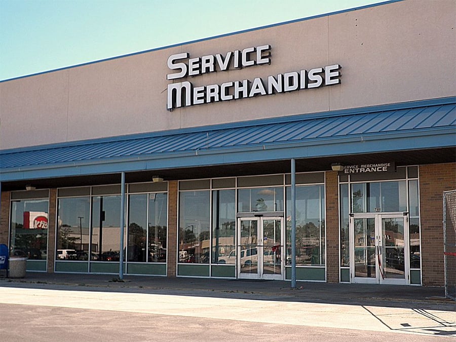 A closed Service Merchandise storefront with large windows, double glass doors, and a sign above the entrance. The building has a blue awning and there are reflections of cars in the glass.