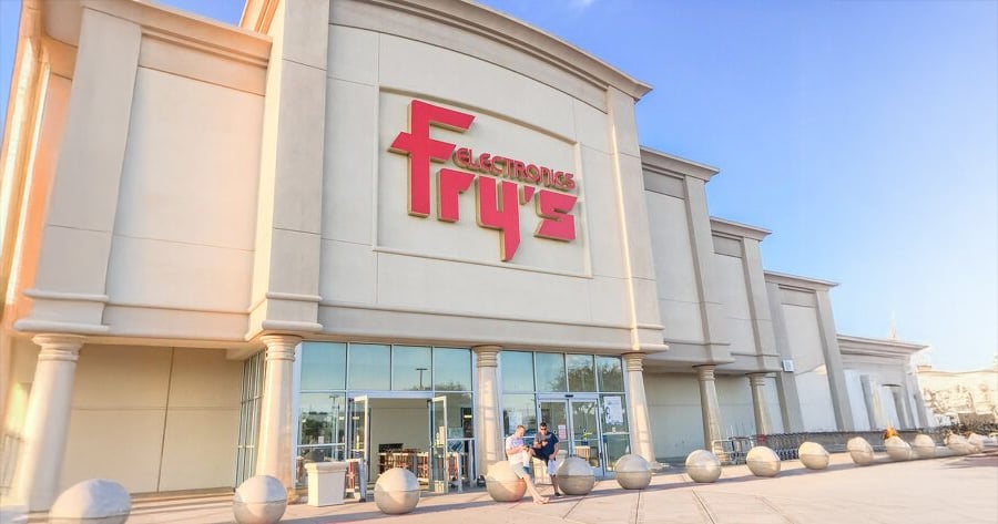 The image shows the exterior of a Fry's Electronics store with large red signage above the entrance and glass doors, under a bright blue sky.