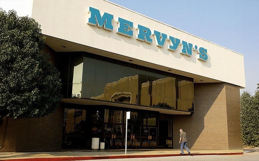 A person walks in front of a large Mervyn's store with a white facade, reflective windows, and teal letters, flanked by trees.