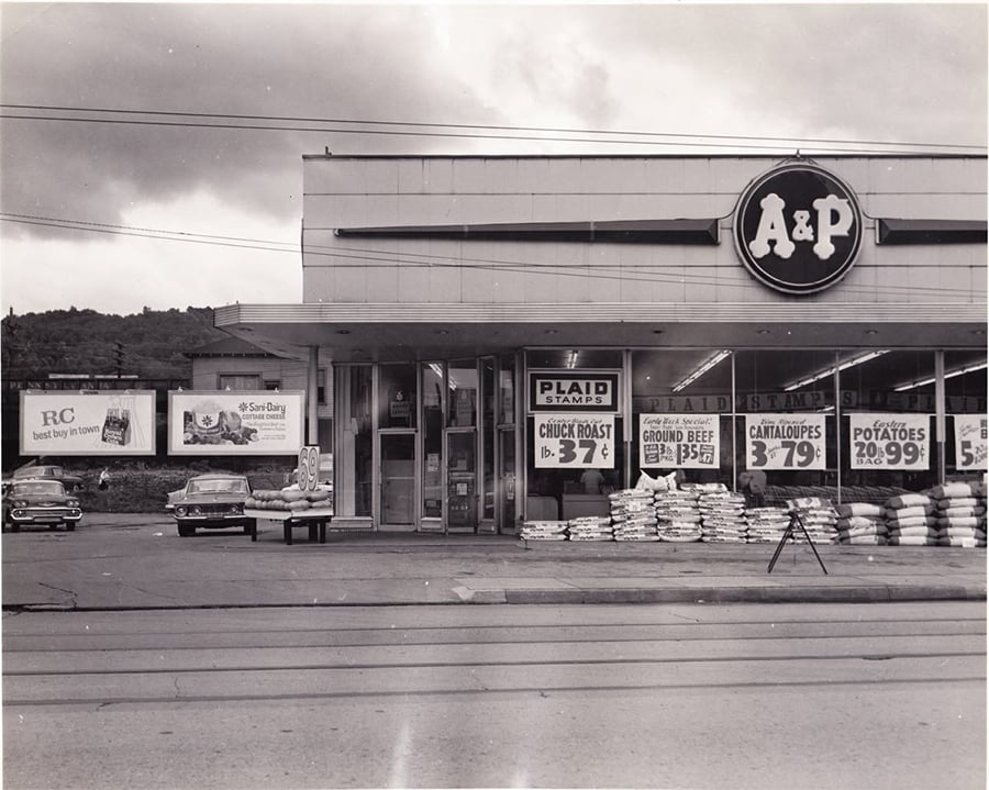 Black and white photo of an A&P grocery store with cars parked outside. Signs advertise sales on chuck roast, ground beef, cantaloupes, and potatoes. Bags are stacked near the entrance. Large A&P logo is above the door.