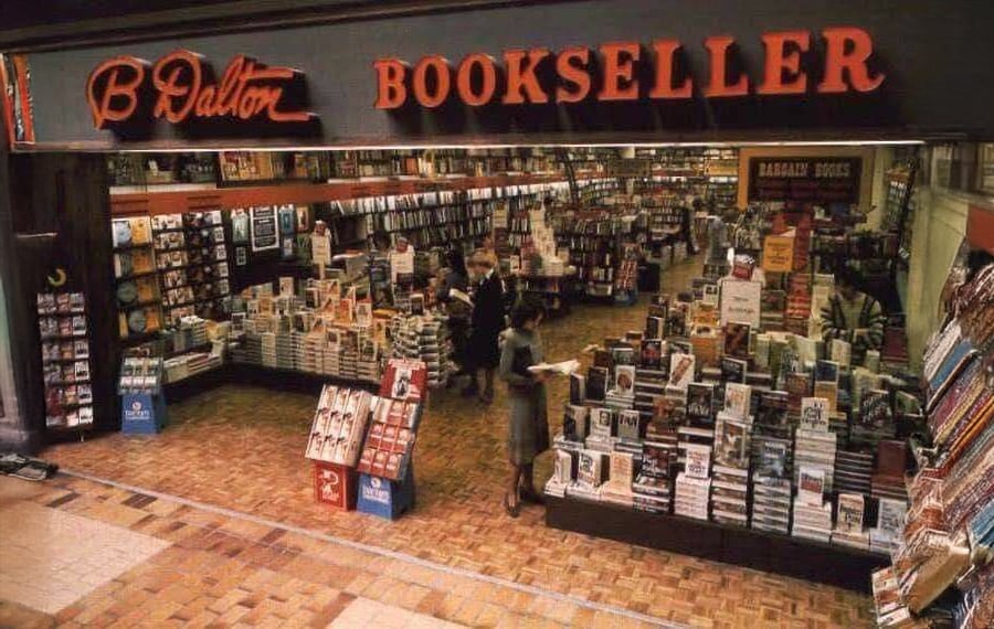 A B. Dalton Bookseller store in a mall, with shelves and tables filled with books and a few people browsing inside. The store's large red sign is visible above the entrance.