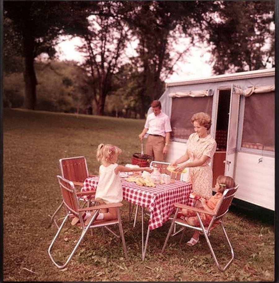 A family of four enjoys a picnic at a table with a red checkered cloth beside a camper; two children sit while the parents prepare food outdoors on a grassy field with trees in the background.