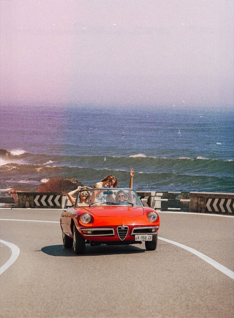 Three people ride in a red convertible, smiling and raising their arms while driving along a coastal road with the ocean and waves in the background.