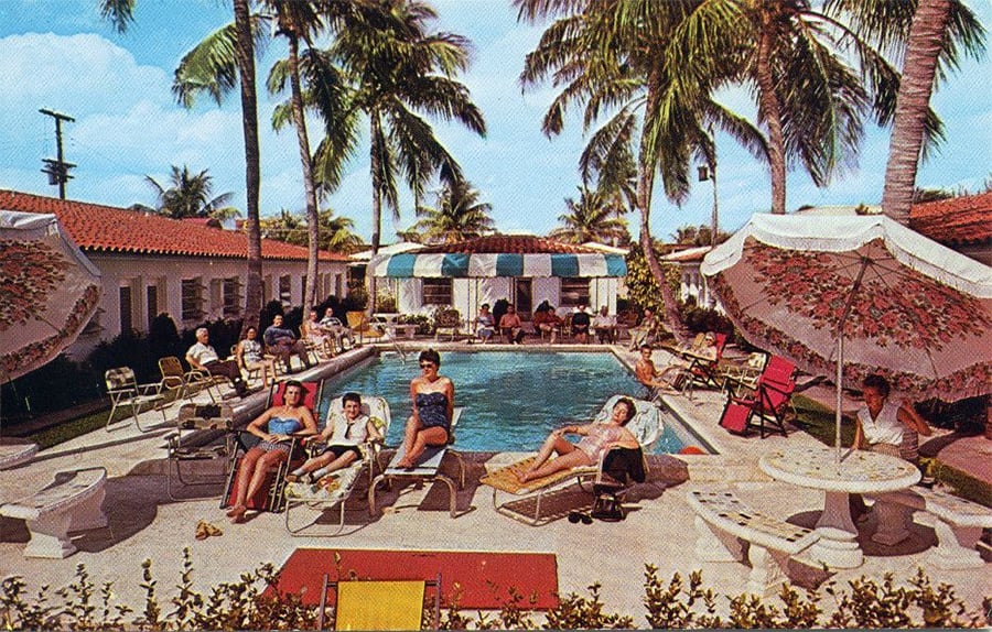 People relax on lounge chairs around a pool at a tropical resort, with palm trees, umbrellas, and a red-roofed building in the background. The scene has a vintage, mid-20th-century vibe.