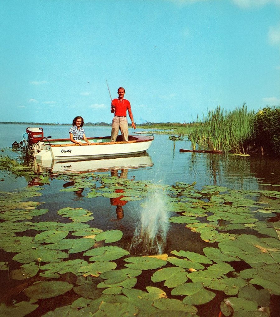 Two people fish from a small boat named “Candy” on a lake covered with lily pads; one person stands holding a fishing rod as water splashes where a fish strikes.