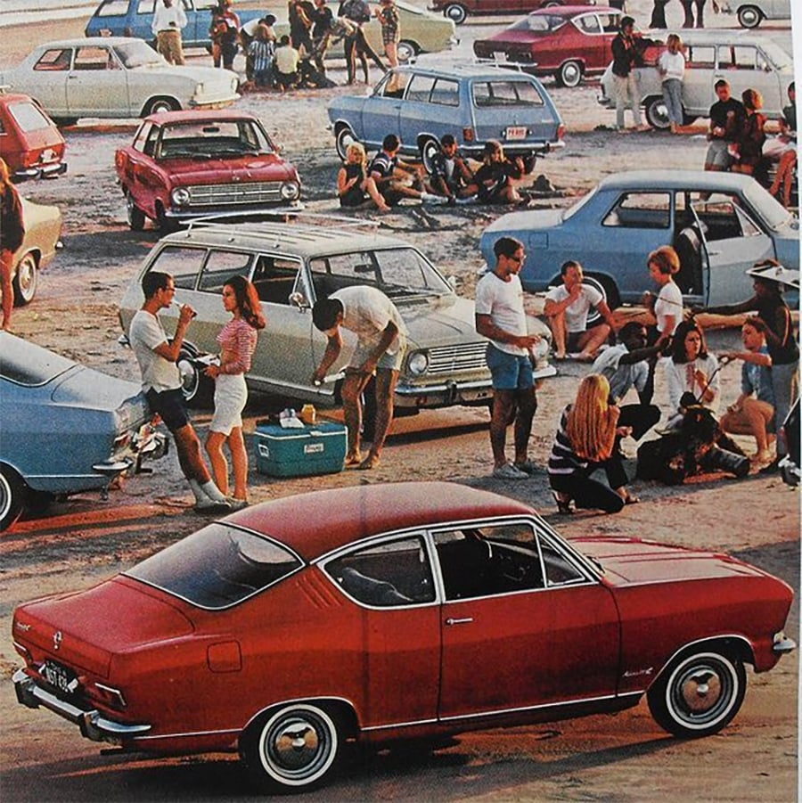 People gather in a parking lot filled with vintage cars, socializing, sitting, and standing in groups. A red coupe is prominent in the foreground. Coolers and casual summer clothing suggest a daytime outdoor event.