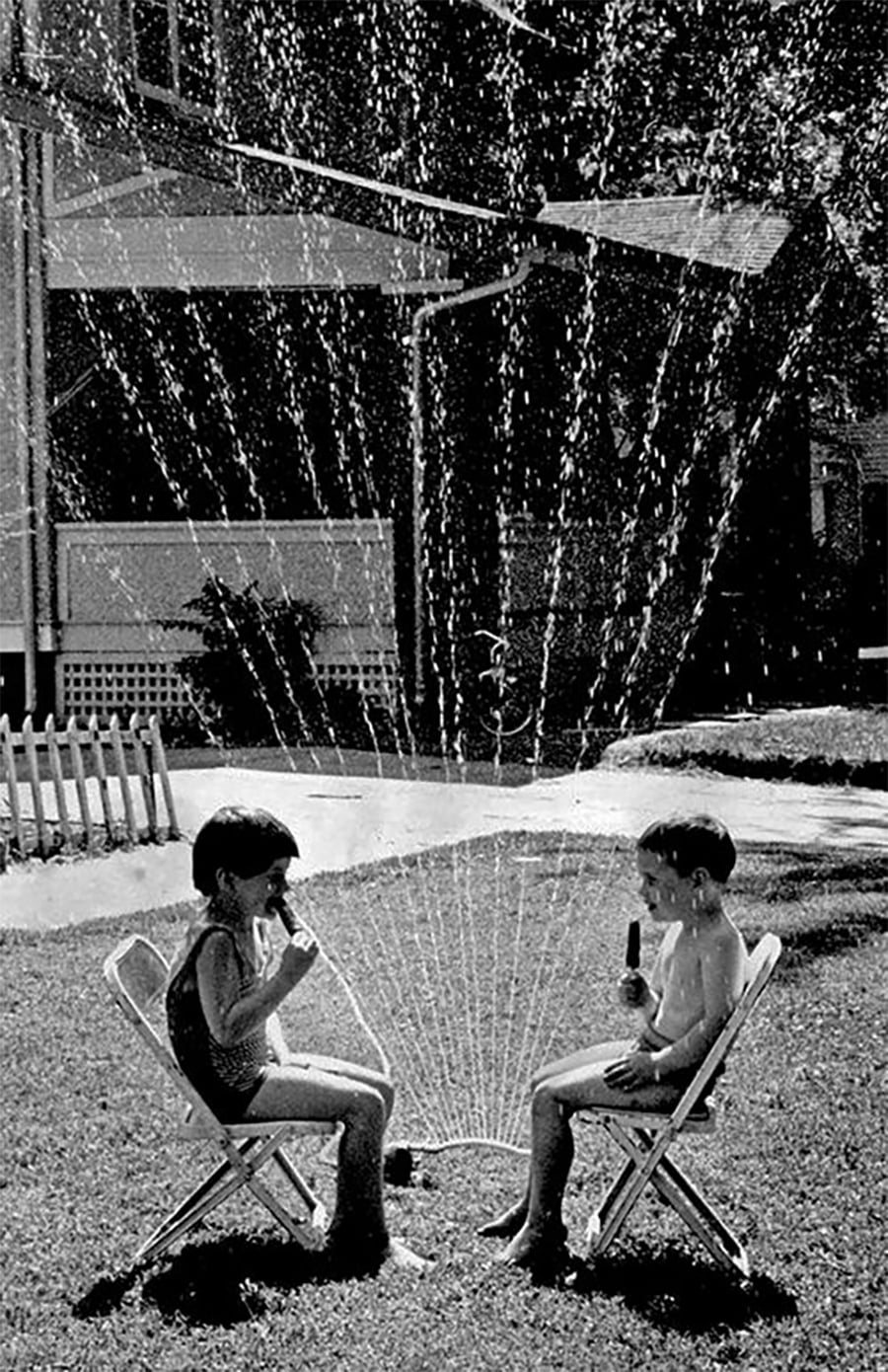Two children sit on chairs, eating popsicles in swimsuits, while a lawn sprinkler sprays water over them on a sunny day. A house and white fence are visible in the background.