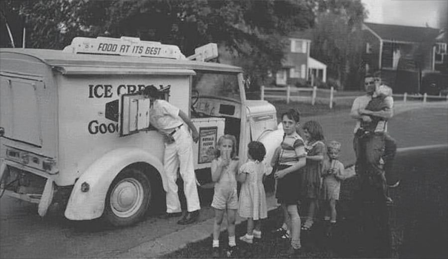 Children line up at an old-fashioned ice cream truck as the vendor serves them, while a man holding a child stands nearby. The scene is set in a suburban neighborhood.