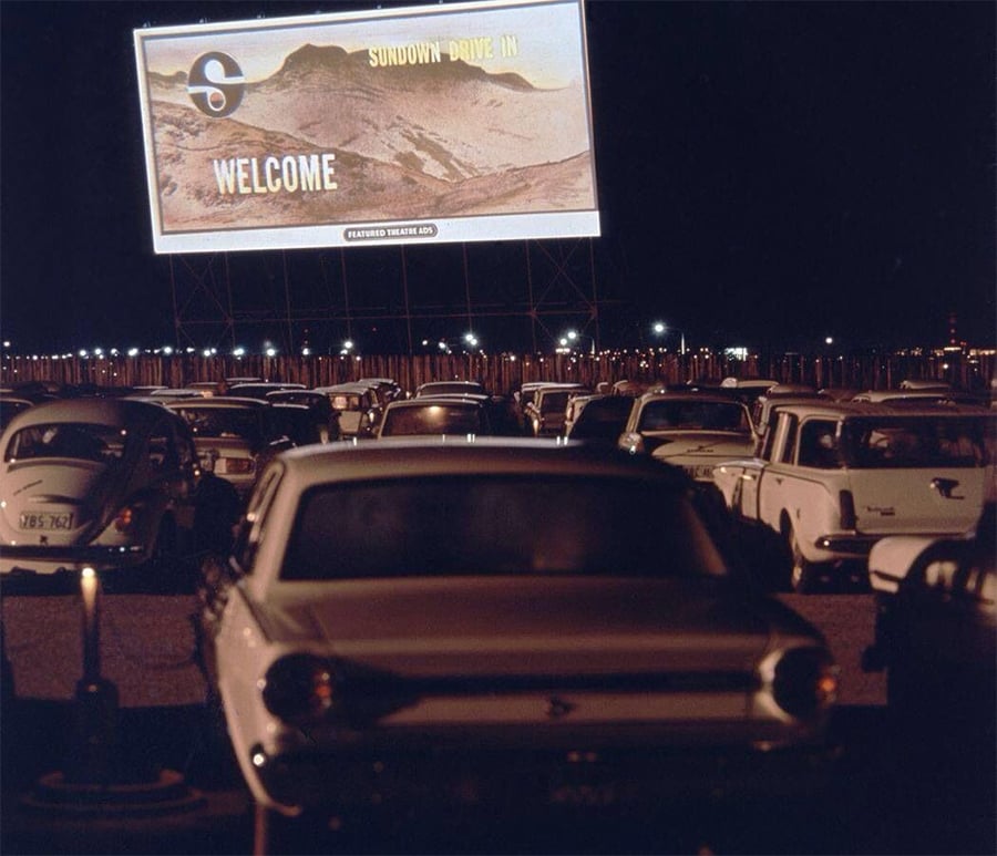 Cars parked at a drive-in theater at night, facing a large outdoor screen displaying “Sundown Drive In Welcome” with a mountain scene. The image has a nostalgic, vintage feel.