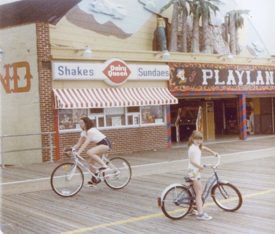 Two kids ride bikes on a boardwalk near a Dairy Queen and an arcade named Playland, with a mural of palm trees and mountains above the shops.