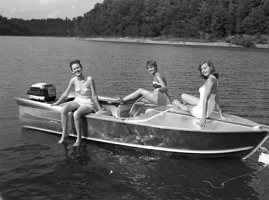 Three women in swimsuits sit and smile on a small metal motorboat floating on a lake, with a forested shoreline in the background.