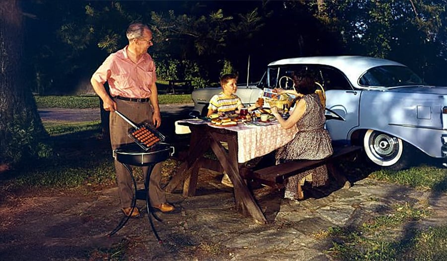 A man grills food beside a picnic table where a woman and child sit, with a classic car parked nearby. They are enjoying an outdoor meal in a park on a sunny day.
