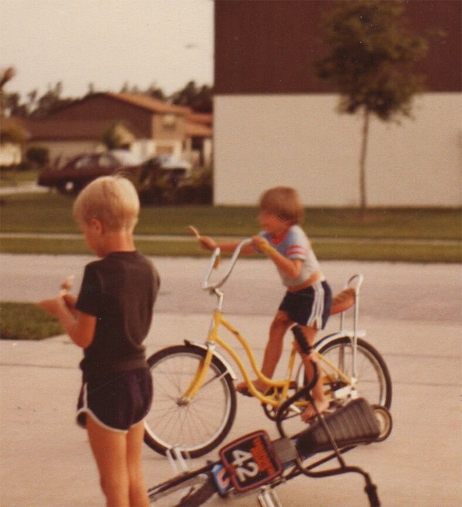 Two young kids play outside; one stands eating ice cream, and the other rides a yellow bike. A blue bike with the number 42 is nearby. Suburban houses and a tree are in the background. The image has a vintage feel.