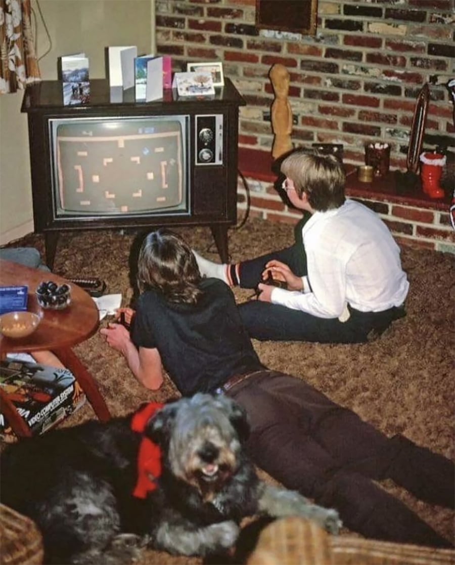 Two kids play a vintage video game on an old TV in a cozy, carpeted living room as a furry dog with a red ribbon lies in the foreground, with greeting cards on the TV and brick walls in the background.