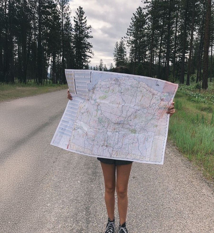 A person stands on a rural road holding a large map that covers their face and upper body, surrounded by trees and greenery under a cloudy sky.