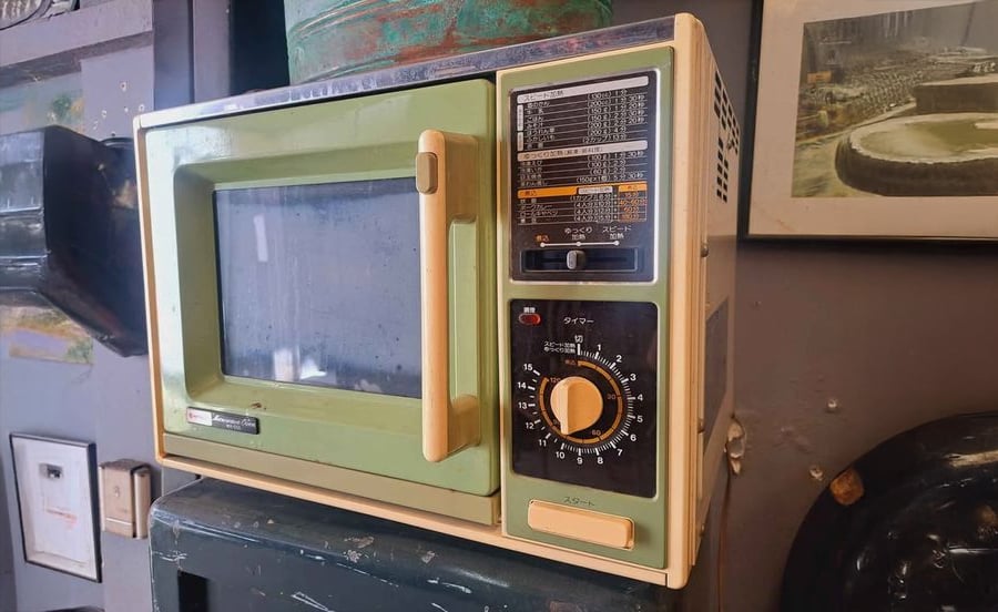 A vintage microwave oven with a green and cream exterior, a clear window, a large dial, and Japanese text on the front panel, sits on a dark surface against a wall with framed pictures.