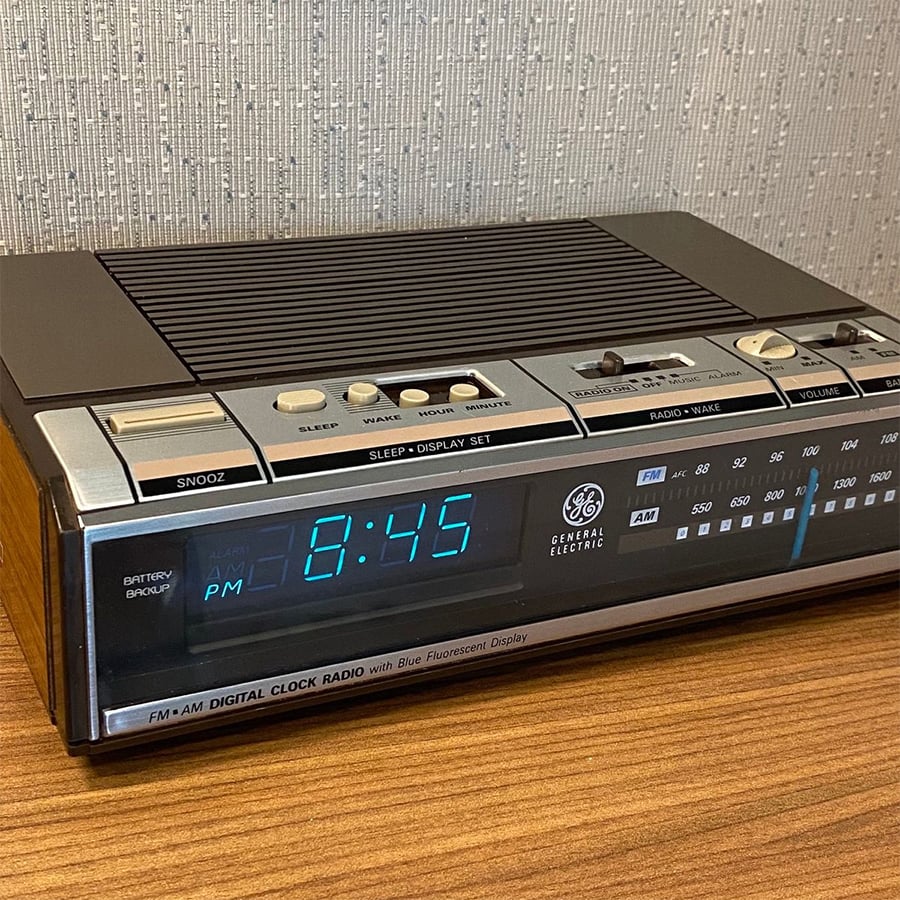 A vintage GE digital clock radio displays 8:45 PM in blue numbers. It has AM/FM dials, several buttons, and sits on a wooden surface against a textured wall.