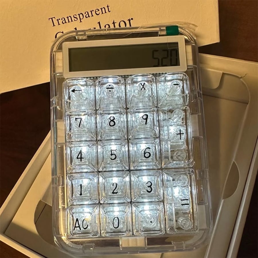 A transparent calculator with illuminated keys displays "520" on its screen. The calculator rests on a table near packaging and a booklet labeled "Transparent Calculator.