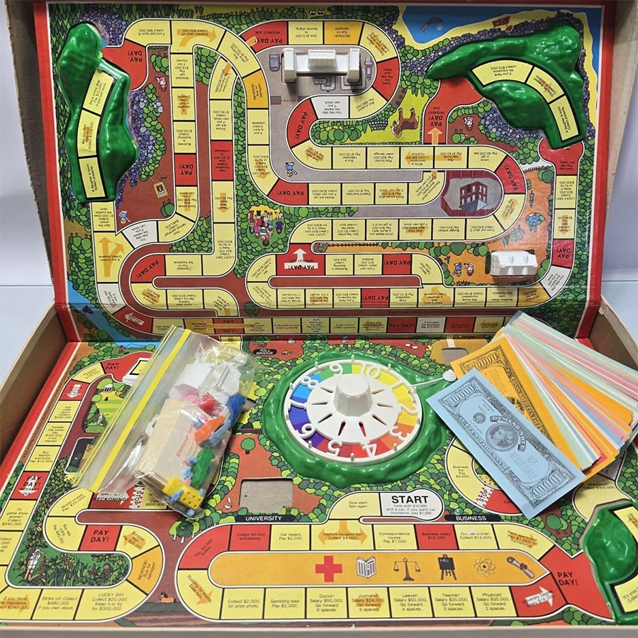 The Game of Life board game is set up, showing the colorful path, spinner, play money, cards, and player pieces in bags, all inside an open game box.