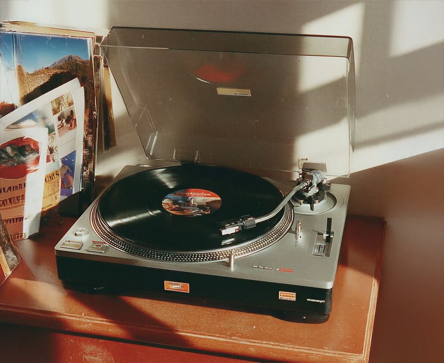 A vintage record player with an open lid spins a black vinyl record on a wooden table, next to several open books or magazines. Sunlight casts shadows across the scene.
