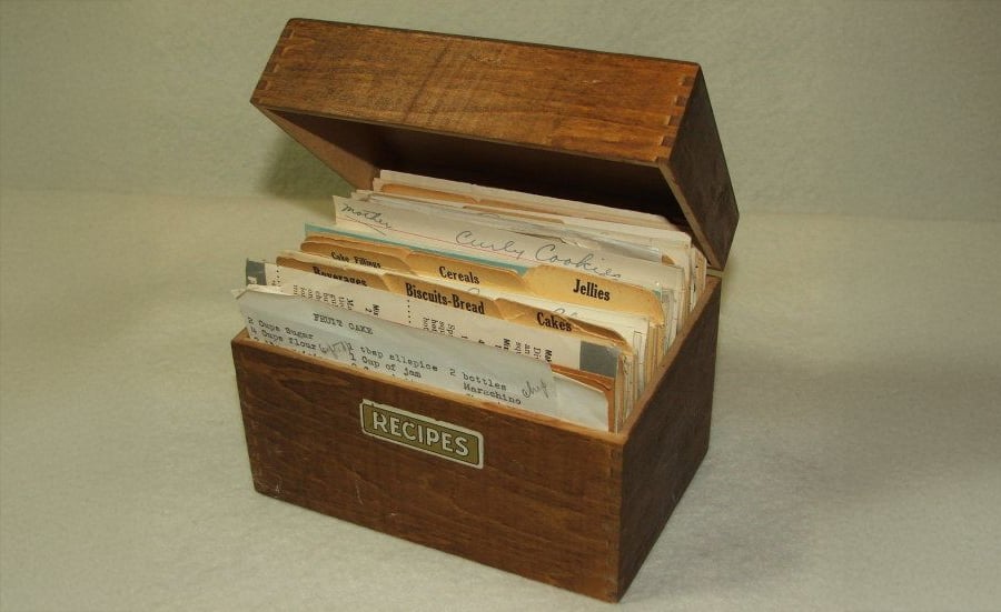 A wooden recipe box with a hinged lid is open, revealing index cards with handwritten and typed recipes. Divider tabs label categories like "Cereals," "Biscuits-Bread," and "Cakes." The box has a "RECIPES" label.