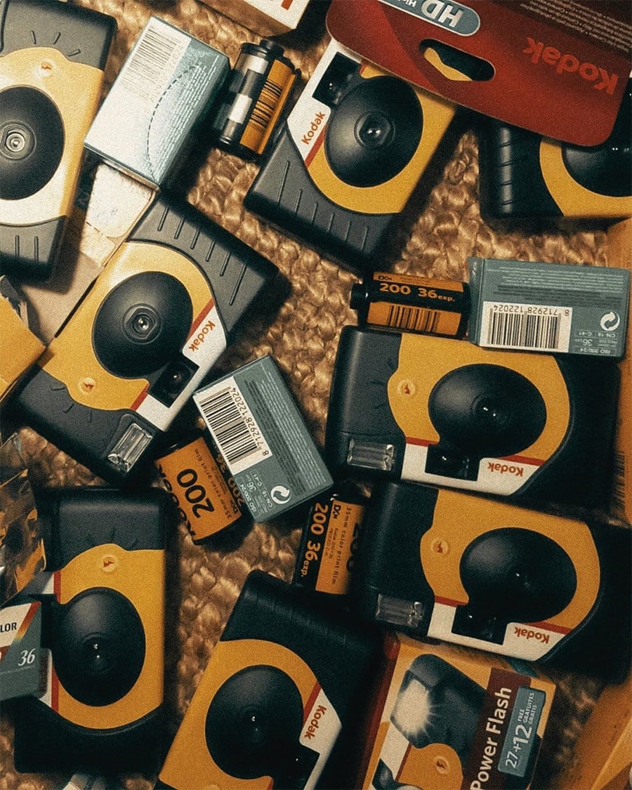 Scattered Kodak disposable cameras and film rolls lying on a woven surface, with visible orange, yellow, and black packaging.