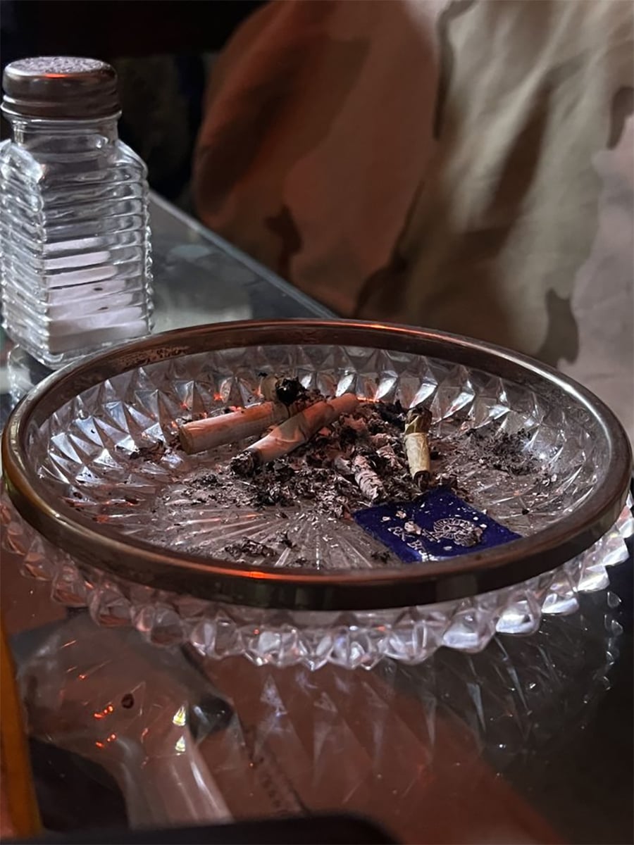 A glass ashtray filled with cigarette butts and ashes sits on a reflective table next to a clear salt shaker. The background shows part of a camouflage-patterned fabric.